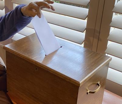 A hand inserts paper into an antique ballot box.