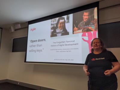 Donna Benjamin stands in front of a screen displaying a screenshot of an article by Shannon Mitchell, published 18 May 2020  called "The forgotten feminist history of agile development" and a quoted snippet saying "Open doors, rather than selling keys" with portraits of Sister Corita Kent, and Octavia Butler. Photo by Fiona Tweedie