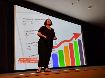 Donna speaking in front of a large screen showing a survey and colourful graph. Photo Credit: Josh Simmons