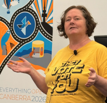 Donna Benjamin wearing the yellow OpenAustralia "They Vote For You" T-shirt in front of the EverythingOpen 2026 Canberra banner